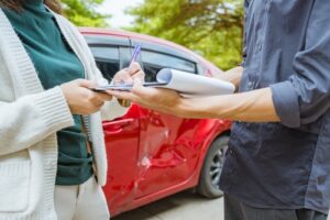 person signing clipboard documents beside damaged vehicle after car accident
